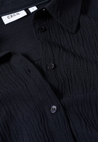 Schwarzes Texturshirt mit einem zerknitterten Stoffdesign, Knopfverschluss und einem Etikett mit der Aufschrift "CECIL" in Größe "S".
