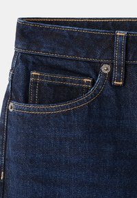 Tmavě modré džínové kalhoty zobrazující přední kapsu, malou kapsičku na mince, poutko na pásek a viditelné žluté švy.