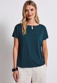 Teal short-sleeve top met een keyhole detail bij de halslijn, gemaakt van zachte stof. Draag het met beige linnen broek, zichtbare gladde textuur.