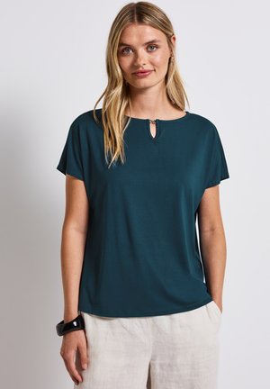 Top corto teal con dettaglio a chiave nel décolleté, realizzato in tessuto morbido. Indossato con pantaloni di lino beige, texture liscia visibile.