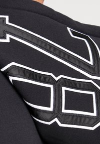 Černá mikina s velkým číslem 10 vyšitém bílou nití a silným bílým okrajem. Hladká textura s atletickými designovými prvky a střihem.