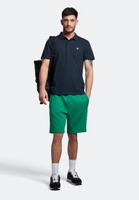 Polo shirt navy con logo giallo, abbinato a pantaloni corti a quadri verdi. Il modello indossa sneakers nere e calzini bianchi alla caviglia, portando uno zaino nero.