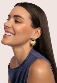 Los pendientes de oro presentan un diseño de explosión estelar con una gema azul redonda en la parte superior. Se llevan con una blusa sin mangas de color azul profundo contra un fondo claro.