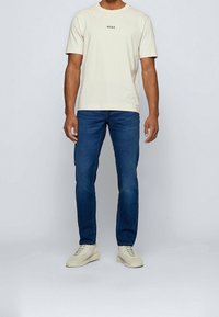 Man i krämfärgad BOSS t-shirt med kort ärm, mörkblå jeans och krämfärgade sneakers, stående mot en enkel ljusgrå bakgrund.