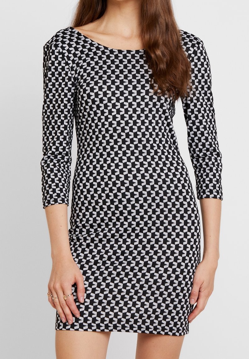 Femme portant une robe ajustée à motifs géométriques noirs et blancs, avec des manches trois-quarts et un col rond.