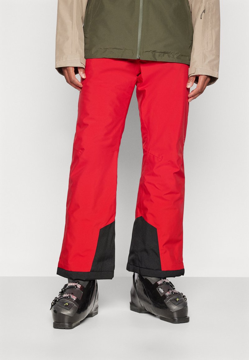 Pantalons de ski imperméables rouges avec des accents noirs, présentant une coupe droite. Associés à des bottes de ski noires mettant en valeur des éléments métalliques.