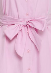 Světlé růžové oblečení na knoflíky s širokým látkovým páskem uvázaným v pěkné mašli v pase.