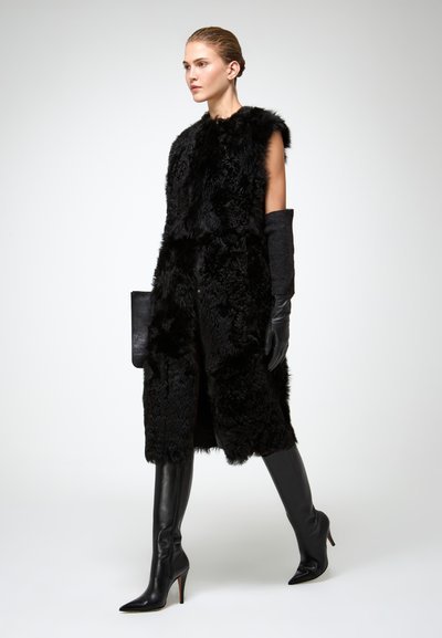 Gilet in pelliccia sintetica nera senza maniche, abbinato a guanti neri e stivaletti con tacco a spillo fino al ginocchio, creando un silhouette elegante.