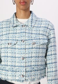 Vrouw in een blauwe en witte gestructureerde tweedjas met zilveren knopen en klepzakken, gedragen over een witte top.