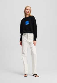 Felpa nera con quadrato grafico blu sul petto, abbinata a pantaloni larghi bianchi e sandali neri con tacco, su uno sfondo neutro.