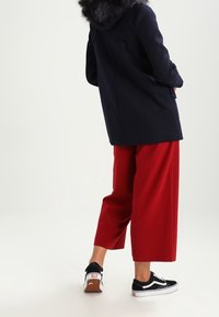 Marinblå rock med huva fodrad med päls, kombinerad med vida röda byxor och svarta sneakers. Har en ren design och texturerat tyg.