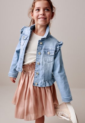 Ung pige smiler og har en lyseblå denimjakke, hvid skjorte, lyserødt skørt og holder en beige sneaker i hælen.