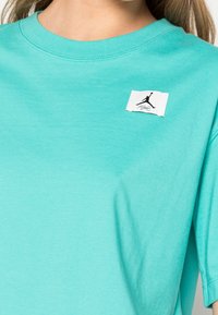 Camiseta de algodón en teal claro con cuello redondo, mangas cortas y una etiqueta blanca con un gráfico de logo negro en el lado izquierdo del pecho.