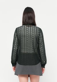 Femme aux cheveux foncés mi-longs, portant un chemisier en dentelle noire à manches longues et une jupe courte grise, debout de dos devant un fond uni.
