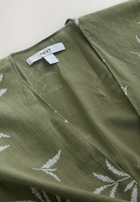 Vêtement vert olive avec des motifs de feuilles blanches et une étiquette de marque "NEXT" blanche cousue dans la zone du col en V.