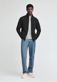 Giacca nera con zip sopra una maglietta grigia, abbinata a jeans azzurri chiari e sneakers beige. Tessuto liscia; vestibilità casual.