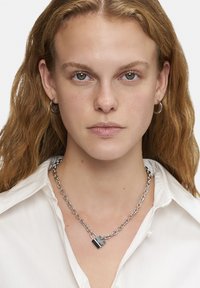 Zilveren ketting met een slotpendant, met een gepolijste textuur en asymmetrisch ontwerp. Gekocht over een wit shirt.