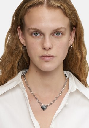 Collier en chaîne argentée avec un pendentif en forme de serrure, au texture polie et au design asymétrique. Porté sur une chemise blanche.