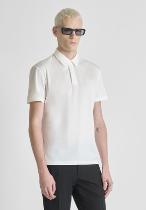 Hombre joven con cabello rubio corto, lleva gafas de sol negras rectangulares, camisa blanca de manga corta con cuello y pantalones negros, posando frente a un fondo liso.