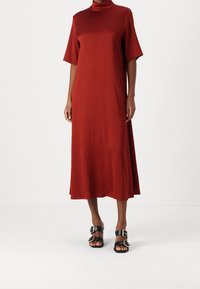 Vestido rojo hasta la rodilla con cuello alto y mangas cortas, hecho de una tela suave. Combinado con sandalias negras con grandes hebillas.