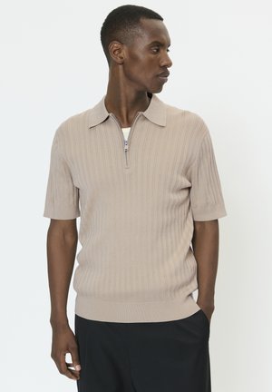 Beige kortærmet polo med ribstrikket tekstur, der har en lynlåslukning ved kraven og en tætsiddende kant. Båret med mørke bukser.