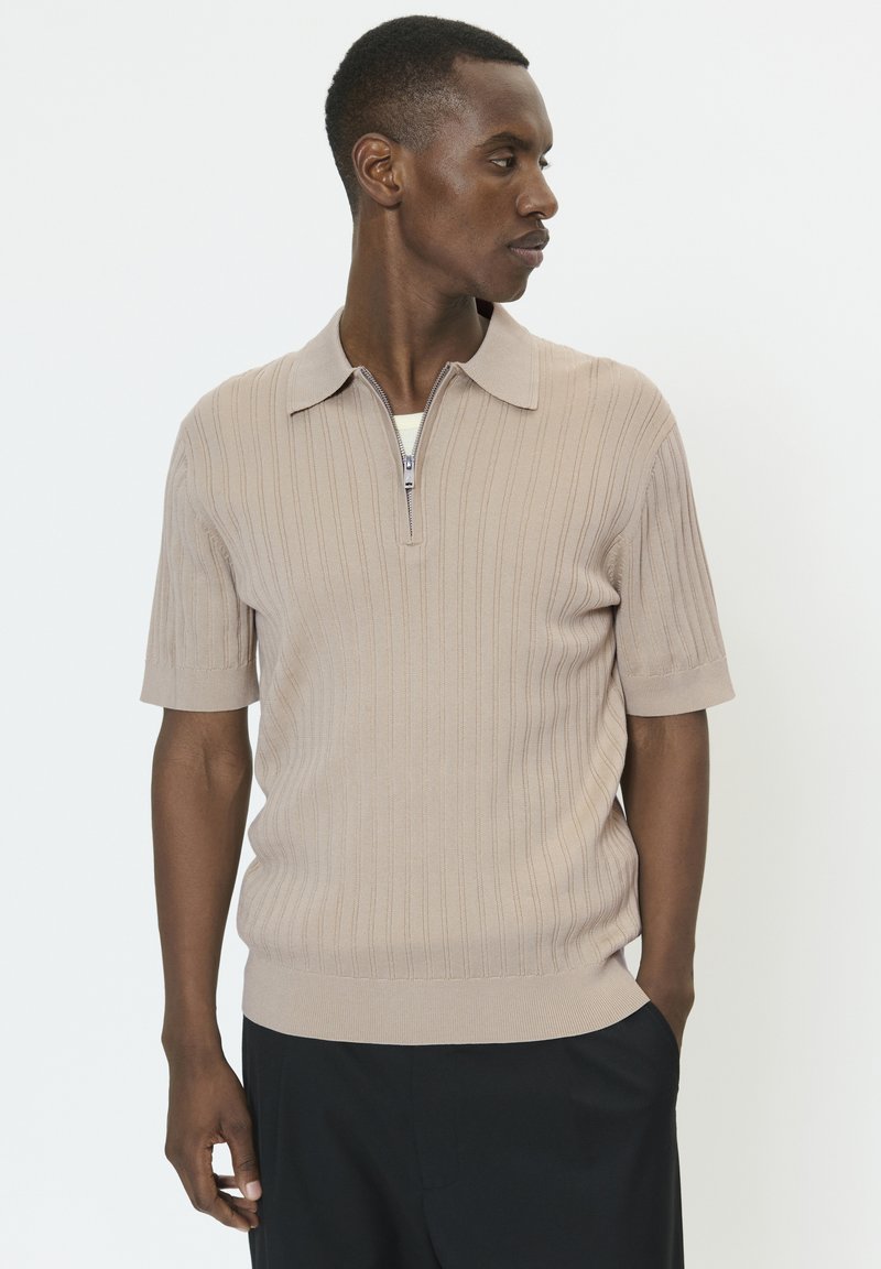 Bež polo majica kratkih rukava s rebrastom teksturom, s ogrlicom na patentni zatvarač i pristajećim rubom. Nošena s tamnim hlačama.
