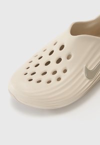 Beige slip-on shoe made of textured material. Features multiple circular cutouts for ventilation and a silver Nike logo accent.