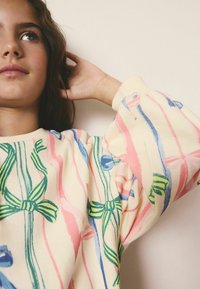 Sweatshirt mit cremefarbenem Grund und bunten Schleifenmustern in Pink, Blau und Grün; hat Raglanärmel und eine weiche Textur.