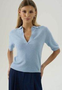 Žena oblečená v svetlomodrej pletenej blúzke s krátkym rukávom a golierom, s tmavomodrými nohavicami, pózujúca s jednou rukou na bok proti jednoduchému pozadiu.