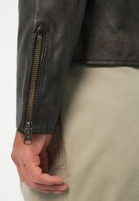 Veste en cuir noir avec un gros plan sur le poignet, mettant en avant une fermeture éclair dorée et la texture du cuir. La manche est légèrement inclinée, révélant une main.