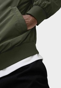 Giacca bomber verde con polsini e vita elasticizzati, dotata di una tasca laterale. Indossata con una camicia bianca e pantaloni neri sotto.