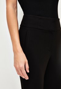 Zwarte op maat gemaakte broek met een gladde textuur, voorzien van een hoge tailleband en zijzakken. Zichtbare plooien versterken het algehele ontwerp.