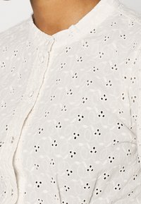 Weiße Bluse aus einem strukturierten Stoff mit runden Ausschnitten und schwarzen Akzenten. Kragenloses Design mit Knopfverschluss.