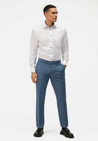 Camicia bianca con bottoni neri e tasca, abbinata a pantaloni leggeri blu con texture. Le scarpe sono nere con suola spessa.
