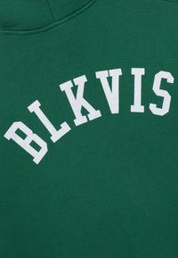 Zelená látka s bílými písmeny ve stylu varsity, která tvoří nápis "BLKVIS" v oblouku, pravděpodobně část mikiny nebo kapucí.
