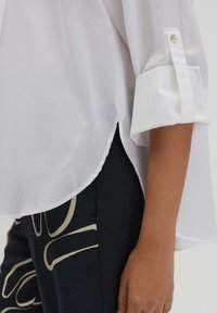 White shirt with rolled sleeves and button detail, featuring a curved hemline. Paired with black pants displaying a light abstract pattern.