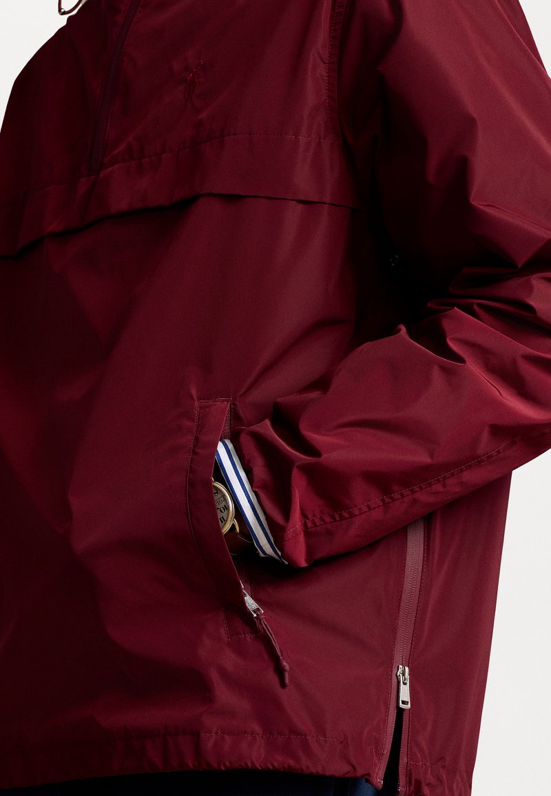 Rote, leichte, wasserdichte Jacke mit einem Halbreißverschluss-Design, elastischen Bündchen und Seitentaschen. Mit einem Logo auf der Brust und kontrastierenden Besätzen.
