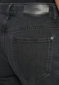 Jeans in denim nero con un'etichetta in pelle sulla vita, cuciture visibili e una tasca posteriore con dettaglio in rivetto metallico.