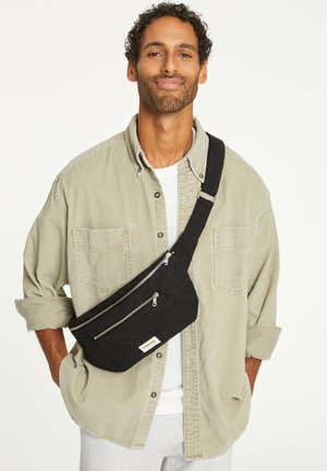 Sacoche en tissu noir avec deux compartiments zippés, une bandoulière réglable et un petit patch logo. Portée en travers du corps sur une chemise légère.