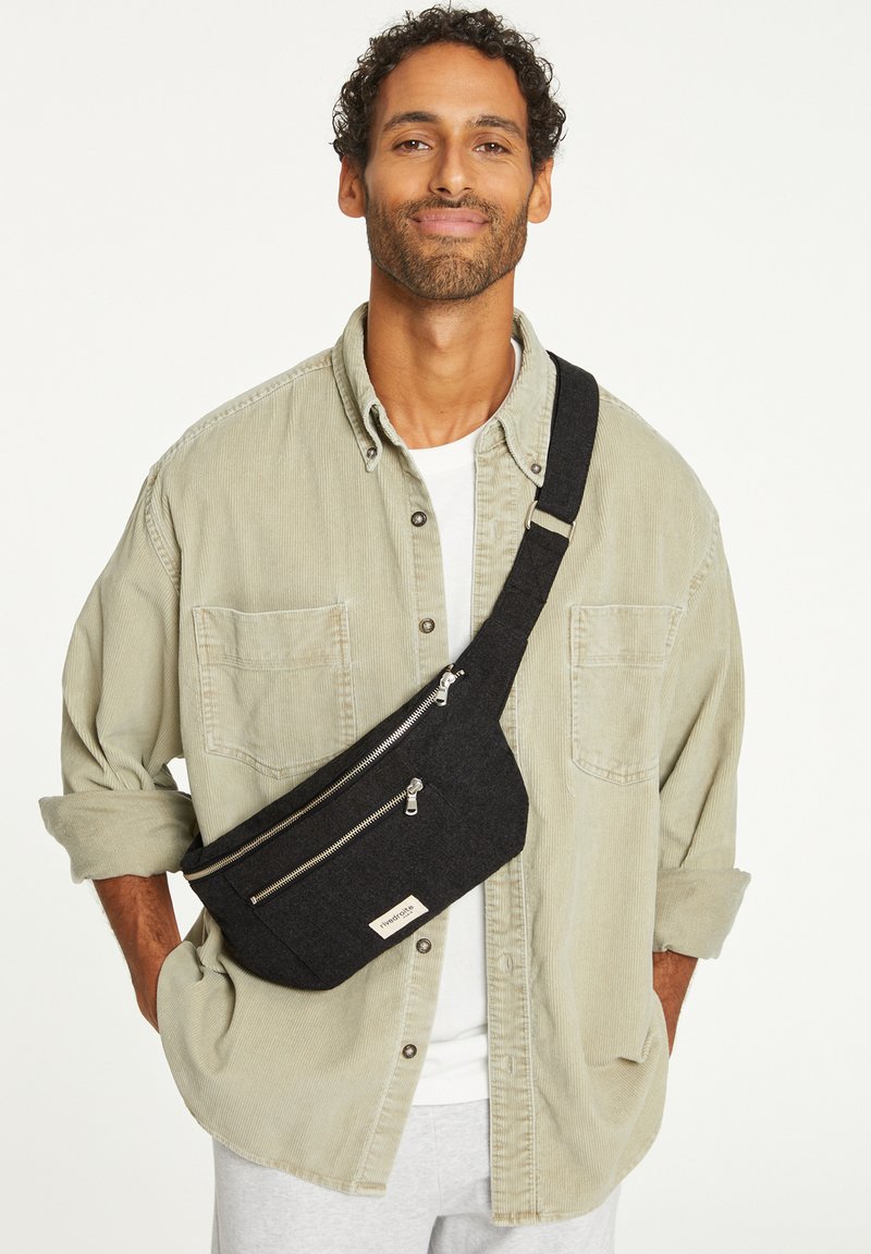 Sacoche en tissu noir avec deux compartiments zippés, une bandoulière réglable et un petit patch logo. Portée en travers du corps sur une chemise légère.