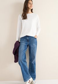 Un haut blanc à manches longues associé à un jean en denim bleu ample. La tenue présente une texture lisse et une veste violette profonde posée sur un bras.