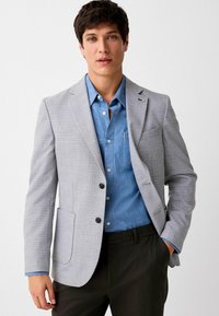 Hellgrauer Blazer mit dezentem Karomuster, zwei Knöpfen und einer Brusttasche, kombiniert mit einem blauen Chambray-Hemd und schwarzen Hosen.