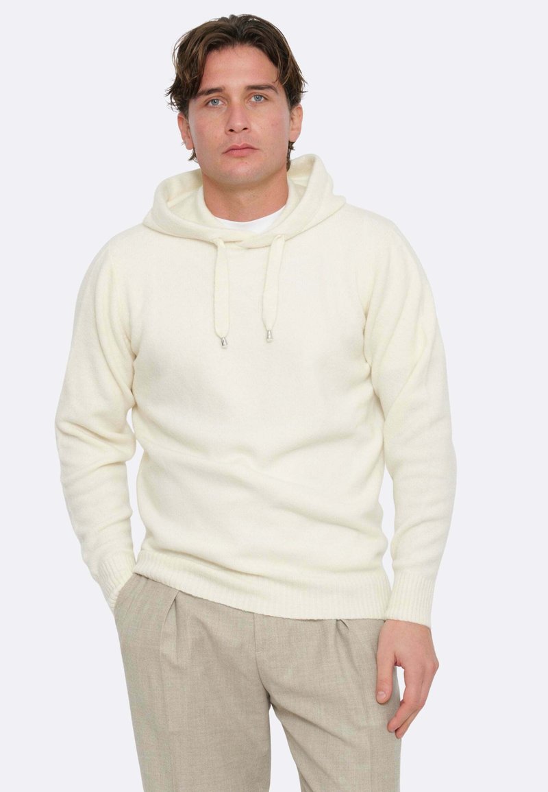 Felpa crema con cappuccio e cordino, con polsini e orlo a coste. Abbinata a pantaloni beige chiaro, che mostrano una vestibilità rilassata e una consistenza morbida.