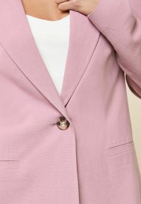 Svetlo roza blejzer iz teksturirane tkanine, z zapiranjem na en gumb in sprednjimi žepi. Ima izrezane reverje in krojen prilegajoč.