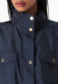 Giacca blu navy con colletto alto, due tasche sul petto, bottoni a pressione e chiusura con zip, realizzata in un materiale liscio e leggero.