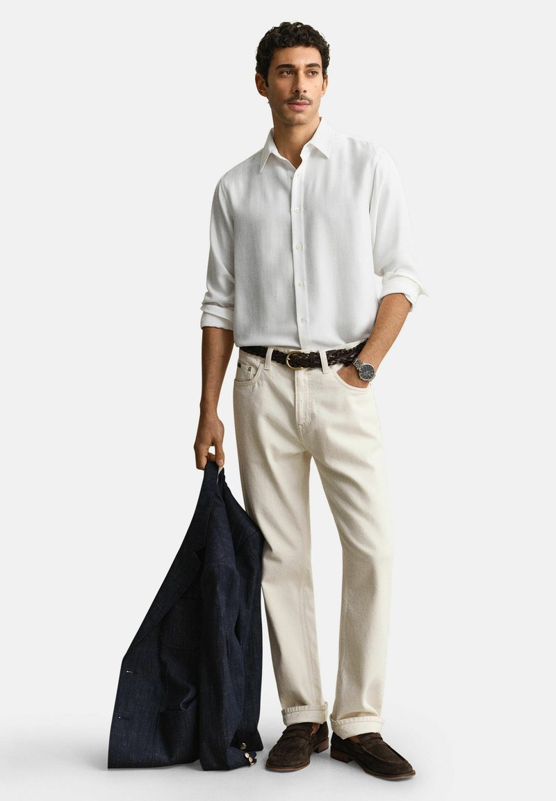 Hombre con camisa blanca abotonada, pantalones crema con cinturón oscuro tejido, mocasines marrones, sosteniendo un blazer azul marino oscuro en una mano.