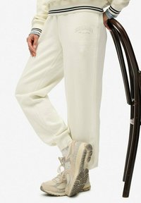 Crème kleurige sweatpants met een elastische tailleband, zijzakken en een geborduurd logoaccent; gecombineerd met beige sportschoenen.