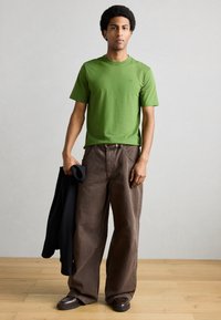 Πράσινο βαμβακερό μπλουζάκι, χαλαρή εφαρμογή, στρογγυλή λαιμόκοψη. Συνδυασμένο με καφέ παντελόνια με φαρδιά μπατζάκια και σκοτεινά παπούτσια, κρατώντας μαύρο σακάκι. Φόντο ξύλινο πάτωμα.