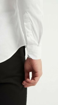 Chemise blanche à manches longues avec des poignets à boutons et un ourlet légèrement courbé. Main placée près d'un pantalon noir, se concentrant sur les détails des manches.