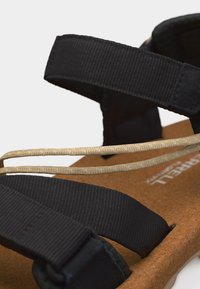 Μαύρο σανδάλι με ρυθμιζόμενους υφασμάτινους ιμάντες και δύο λεπτά μπεζ κορδόνια πάνω σε καφέ φελλό.